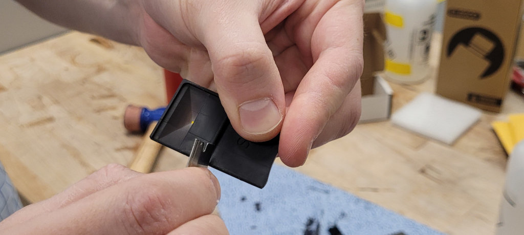 A hand holding a single Fly Pyramid while another hand scratches a razor blade along one of the edges.