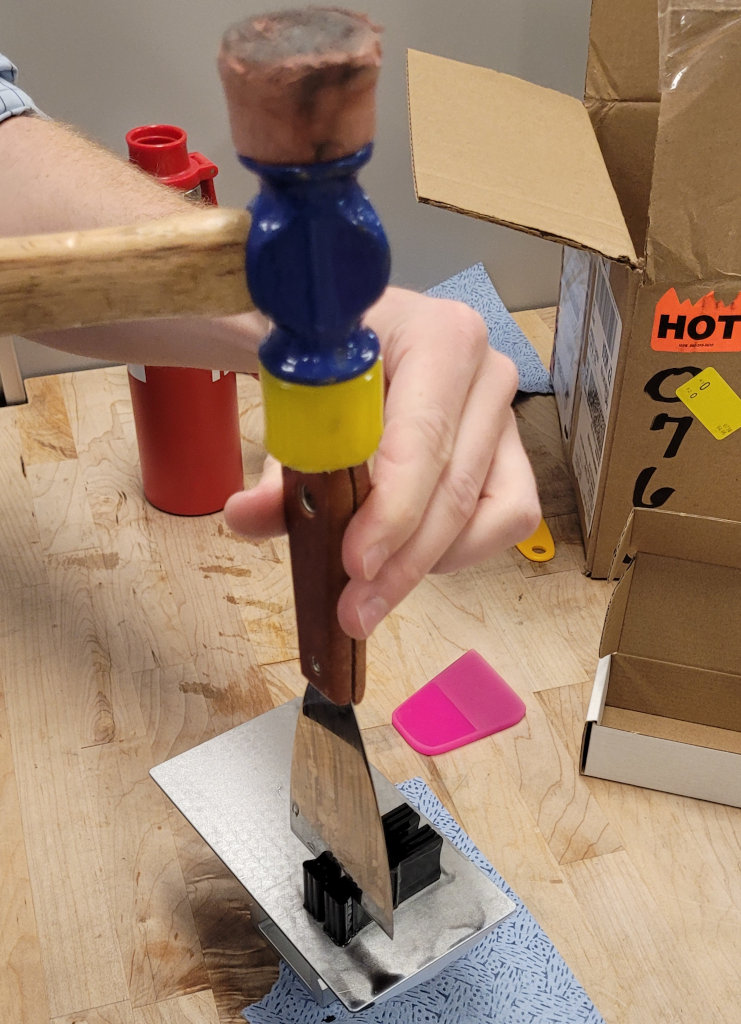 A metal scraper sits on top of the connection between the support and Fly Pyramid, still attached to the build plate. A hammer on top of the scraper handle shows how to separate support and Fly Pyramid.